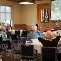 Two women stand in front of a presentation that says "Grand Rapids Ghost Stories"; one holds a microphone and is speaking to seated people